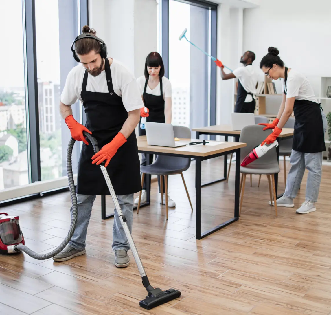 Four people in aprons and gloves, from one of the top office cleaning companies, expertly clean an office. They use vacuums and tools on the wooden floor, meticulously navigating around floor-to-ceiling windows and sleek office furniture.